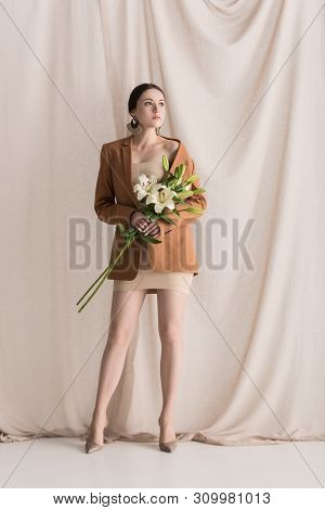 Trendy Model In Beige Dress And Blazer Standing On Curtain Background With Flowers