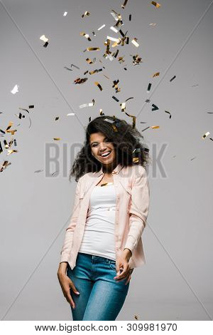 Young African American Woman Smiling Near Falling Confetti While Standing On Grey