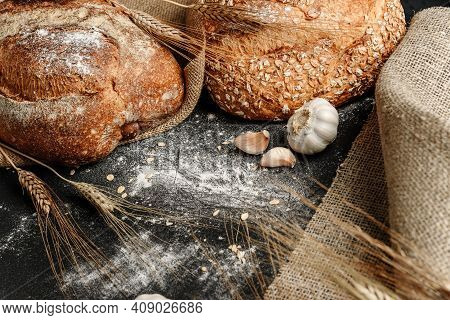 Two Loaves Of Rye Bread On The Table With Ears Of Wheat And Pieces Of Coarse Cloth. Homemade Baking 