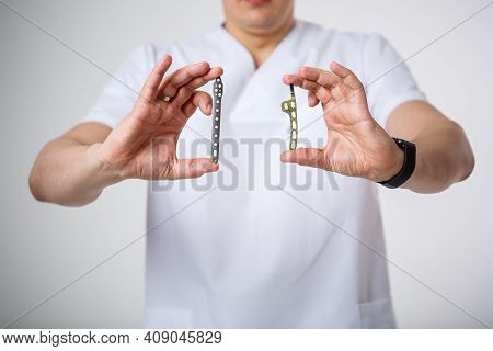 Young Male Doctor In White Surgical Suit Holds And Examines Titanium Plates For Bone Fracture Treatm