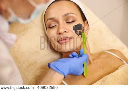 Closeup Of Beautiful Woman's Face And Aesthetician's Hands Applying A Black Beauty Product On Her Fa