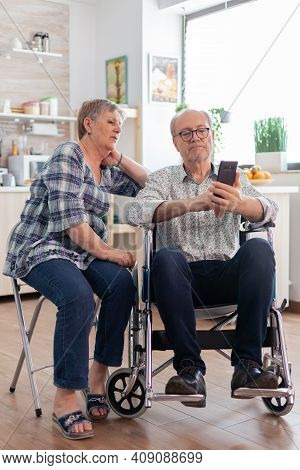 Senior Woman And Disabled Husband In Wheelchair Surfing On Internet Using Smartphone In Kitchen. Par