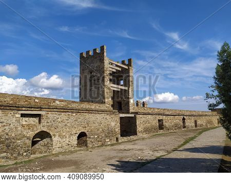 Part Of The Fortress Wall With A Tower In The City Of Sudak In The Crimea In The Summer Against A Bl