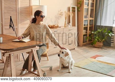 Warm Toned Full Length Portrait Of Modern Blind Woman Sitting At Table In Cozy Home Interior And Pet