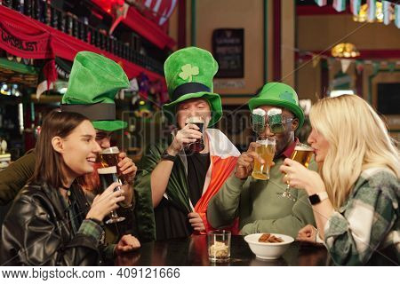 Group of friends in green hats sitting at the table drinking beer and celebrating Patricks Day in the bar