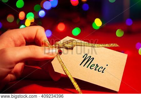Mersi - Text On The Label In A Womans Hand, Close-up, On A Blurred Festive Background.
