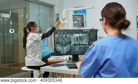 Nurse Analysing Digital X-ray Sitting In Front Of Pc In Stomatologic Clinic, While Doctor With Face 