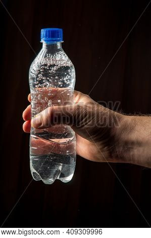 Person Hand Holds A Bottle Of Mineral Water To Quench Thirst. Vertical Image On Dark Background
