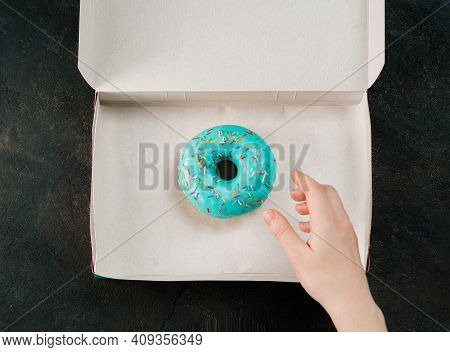Hand Reaches For Donut In Box. One Blue Glazed Donut In Open Paper Box Over Black Textured Backgroun