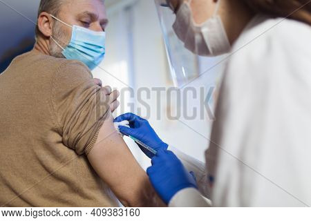 Young Woman Nurse With Surgical Mask And Face Shild Giving Injection To Senior Man. Covid-19 Or Coro