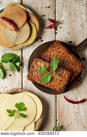 high angle view of some slices of vegan cheese, some tofu fillets and some cold cuts made of tofu in different wooden plates, on a rustic wooden table sprinkled with different herbs and spices
