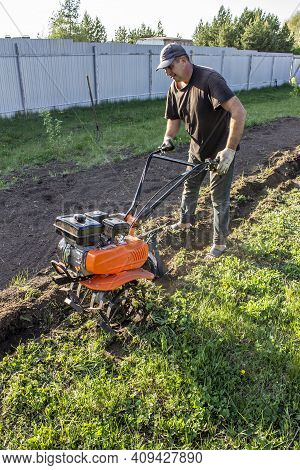 Male In The Spring Plows The Earth In Working Clothes With A Motoblock, Walk-behind Tractor