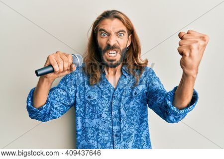 Attractive man with long hair and beard singing song using microphone annoyed and frustrated shouting with anger, yelling crazy with anger and hand raised 