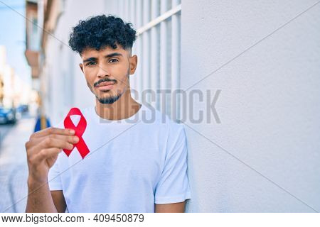 Young arab man with serious expression holding hiv awaraness red ribbon leaning on the wall.