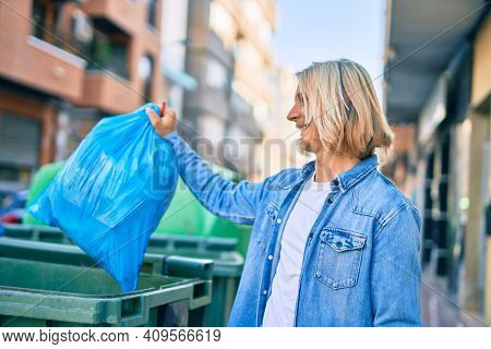 Young blond scandinavian man throwing waste bag to the container at the city.