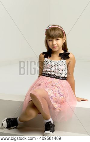 Stylish Girl Sitting On White Staircase. Girl Wearing Nice Dress And Sneakers Sitting On Step Leanin