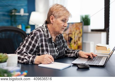 Mature Entrepreneur Taking Notes On Notebook Working In Home Office. Elderly Woman In Home Living Ro