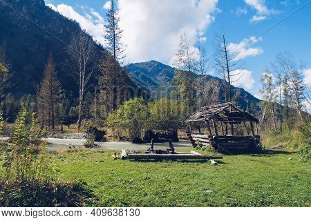 Camping Place With Old Log House By The River Flowing In The Forest Among Trees And Green Lush Bushe