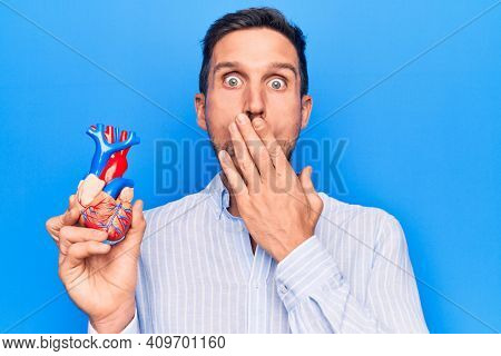 Young handsome man holding heart organ with veins and arteries over blule background covering mouth with hand, shocked and afraid for mistake. Surprised expression