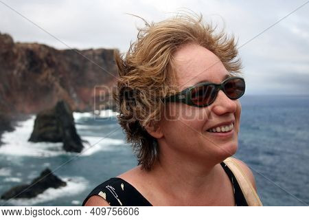Middle Aged Woman Enjoying Wind On The Sea Coast. Woman Is Wearing Sunglesses And Summer Clothes. Po