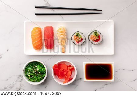 Nigiri sushi and maki roll served on a white plate, seaweed salad, ginger and soy sauce on the side on a stone background. Traditional Asian food, top view.