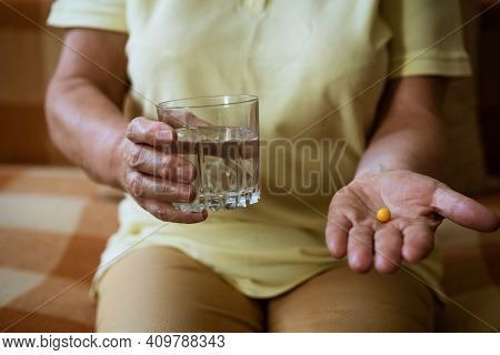 Mature Woman With A Glass Of Water Holds A Tablet In Her Hand While Sitting On The Couch. Treatment 
