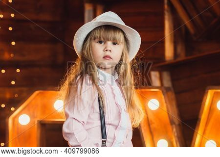 Portrait Of A Little Girl Wearing A Hat On A Background Of Wooden Letters Decorated With Lamps