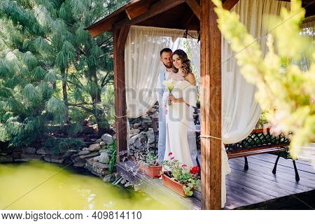 Groom In Blue Suit And Bride With Long Curls With Bouquet In A Gazebo