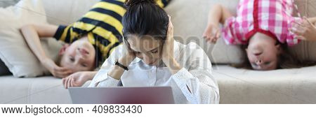 Woman Sits On Floor With A Laptop And Covers Her Ears With Her Hands. Behind Her On Couch Are Childr