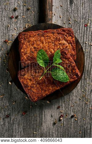 high angle view of some cooked tofu fillets in a round dark wooden tray, on a gray rustic wooden table sprinkled with different spices and herbs