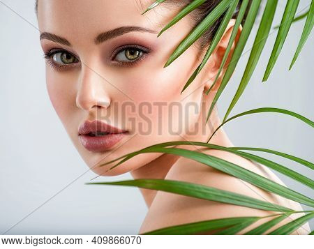 Young beautiful woman with healthy skin of face and palm leaves. Closeup fresh face of an attractive caucasian girl with green plants. Model with bright brown eye makeup. Skin care concept. 