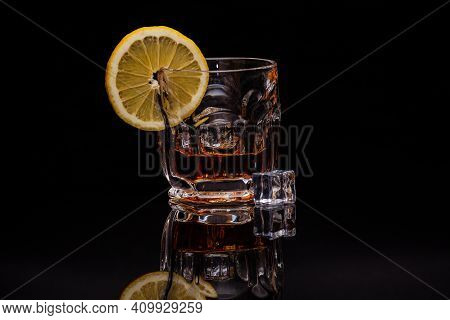 A Glass Of Whiskey With Pieces Of Ice And Lemon Stands On A Black Glass In Which Its Reflection Is V