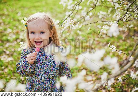 Adorable Little Girl Enjoying Nice And Sunny Spring Day Near Apple Tree In Full Bloom. Outdoor Sprin