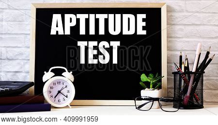 The Word Aptitude Test Is Written On The Chalkboard Next To The White Alarm Clock, Glasses, Potted P