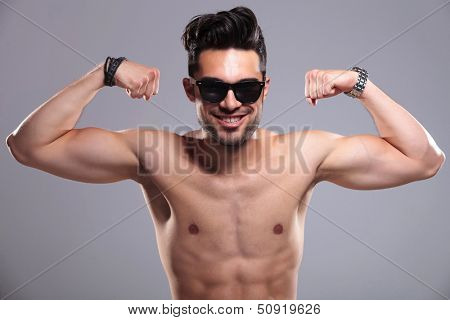 topless young man smiling for the camera while bragging with his arm muscles. on gray background