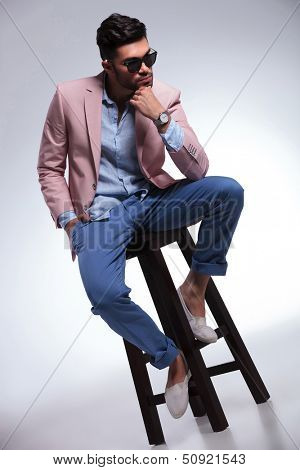 full length picture of a young casual man sitting on a chair and holding his hand at his chin while looking down. on gray background