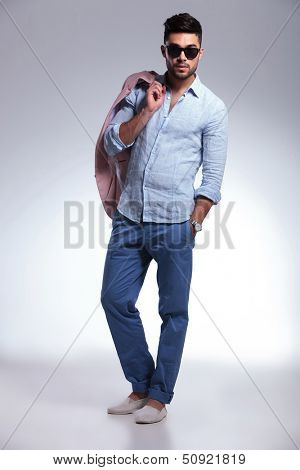 full length photo of a young casual man standing with his jacket on his shoulder and his hand in his pocket while looking into the camera. on gray background