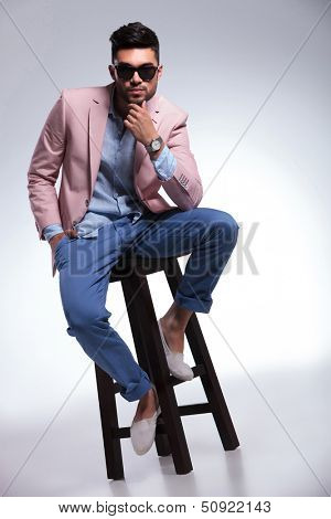 full length photo of a young casual man sitting on a chair and looking into the camera with his hand at his chin. on gray background