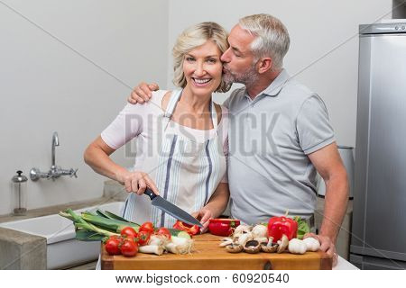 Mature man kissing woman as she chops vegetables in the kitchen at home
