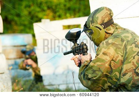 Two paintball sport players in prootective uniform and mask aiming and shoting with gun outdoors