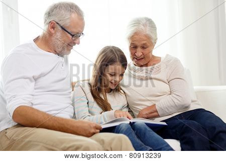 family, generation and people concept - smiling grandfather, granddaughter and grandmother with book sitting on couch at home