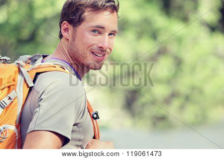Young smiling backpack man in summer forest nature. Happy handsome male adult student looking at camera walking hiking in forest background. School bag or backpacking travel concept.