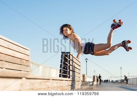Happy young joyful woman doing a balancing act wearing rollerskates. Young woman in skates jumping with the support of a bench. Beautiful young happy woman wearing roller blades doing a stunt. 