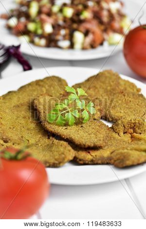 some breaded fillets of seitan in a white ceramic plate, placed on a white table with a lentil salad in the background