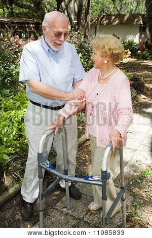Senior couple outdoors.  She's in a walker and he's helping her.