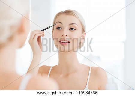 beauty, make up, cosmetics, morning and people concept - young woman applying makeup drawing eyebrow with brush and looking to mirror at home bathroom