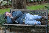 A full view of a homeless man asleep on a park bench with his wine bottle.
