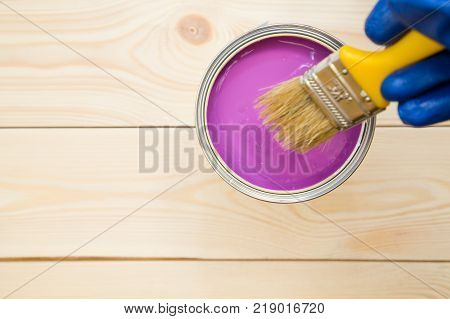 Man's hand in blue glove. A tin can of fuschsia oil paint with a brush on a light uncolored wooden background. Close up. Top view. Space for your text or pruduct display.
