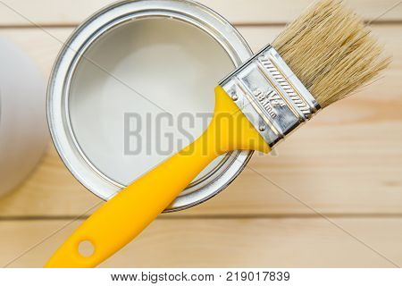 Repair in a house. Tin can of white oil paint with a yellow brush and can of white paint on a light uncolored wooden background. Close up. Top view. Space for your text or pruduct display.