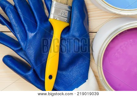 Protection for redecoration work. A pair of blue latex gloves, a brush and tin cans of oil paint on a light uncolored wooden background. Close up. Top view. Space for your text or pruduct display.